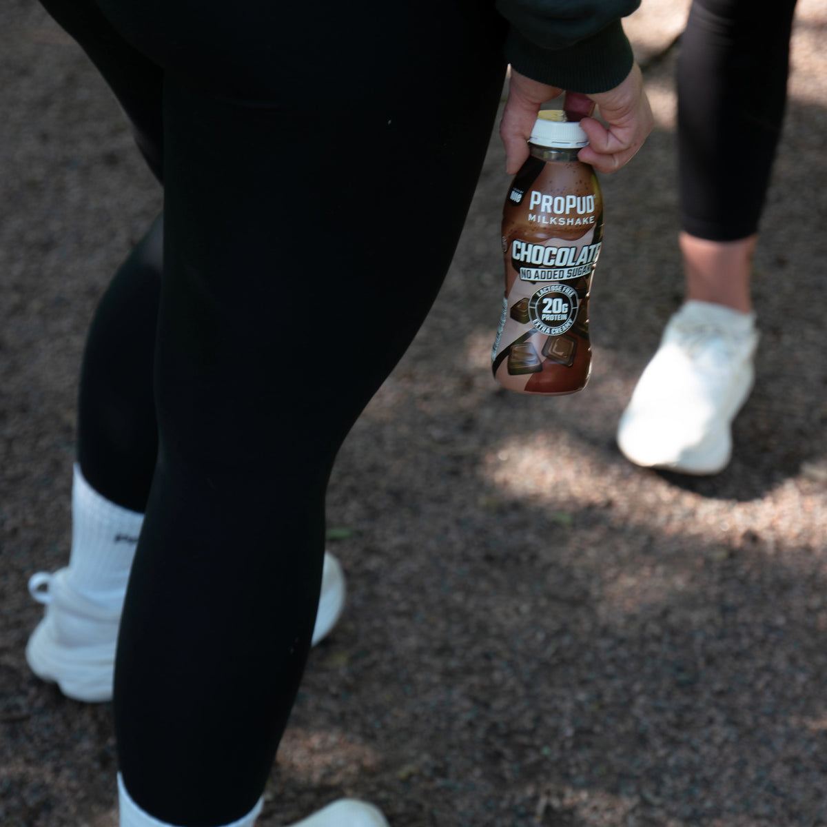 A person outdoors wearing black leggings and white sneakers while holding a bottle of Propub Chocolate Energy Drink. The background shows a gravel path with dappled sunlight and the legs of another person in white sneakers.