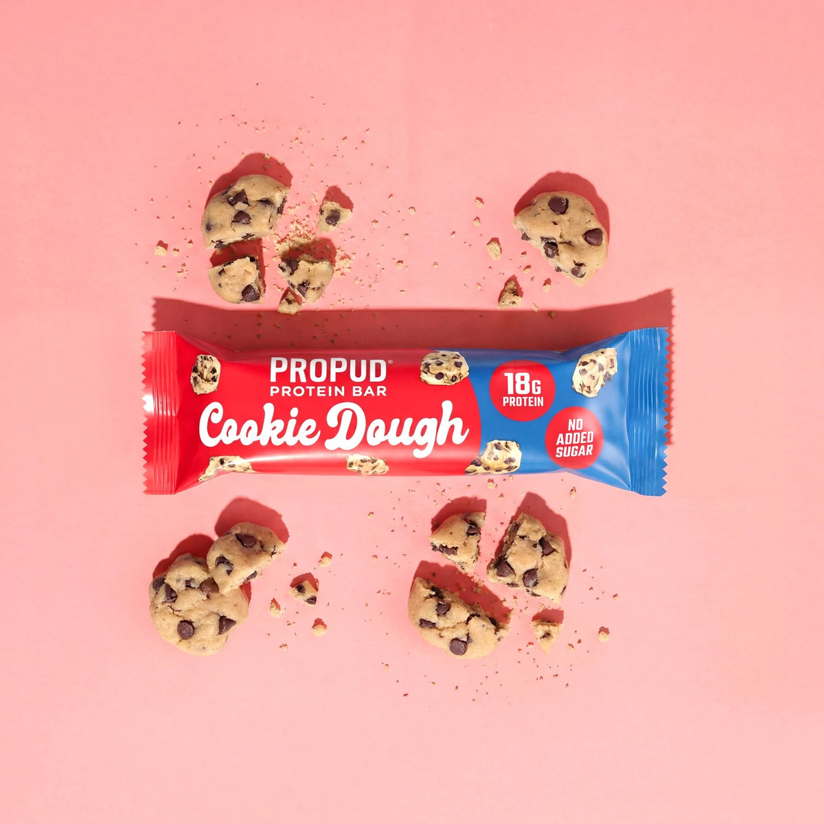 A wrapped Propud Protein Bar in Cookie Dough flavor, laid horizontally on a solid pink background with several broken chocolate chip cookies and red frosting pieces arranged around it.