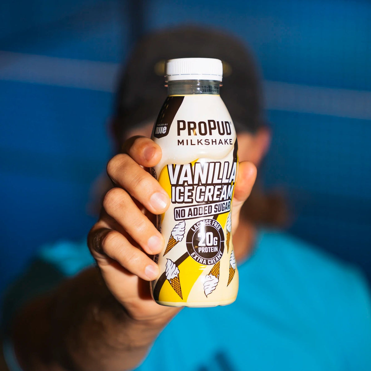 A person in a blue Adidas shirt holds a bottle of Propudo Milkshake Vanilla Ice Cream toward the camera against a blurred blue background.