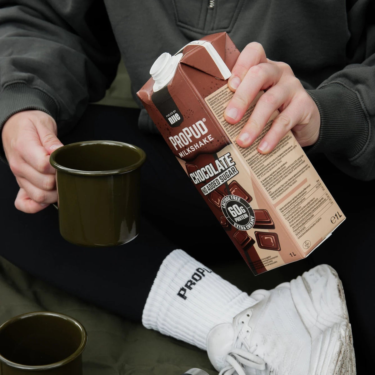 A person in a dark gray hoodie and white socks holds a carton of Propud Chocolate Milkshake while pouring it into an olive green mug. A matching Propud stainless steel bottle and a chocolate bar are visible on a dark green surface.
