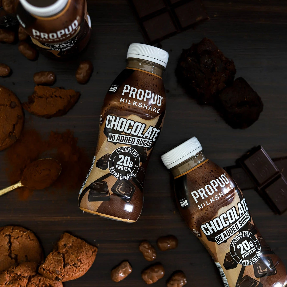 Two bottles of Propudo Milkshake Chocolate Milkshake with chocolate cookies, chocolate chunks, and dark chocolate bars arranged on a dark wooden surface.